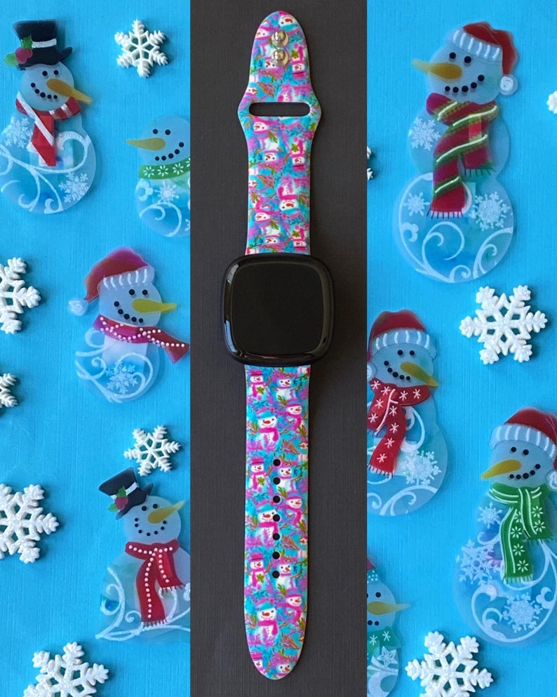K&ouml;nnte beinhalten: Ein pinkfarbenes, blaues und wei&szlig;es Armband mit einem schwarzen Zifferblatt. Das Armband hat ein sich wiederholendes Muster aus Schneem&auml;nnern.
