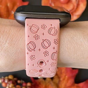 May include: A pink smartwatch band featuring a repeating pattern of pumpkins, flowers, and stars. The band is attached to a dark gray smartwatch face. The design is a seasonal, fall-themed accessory.