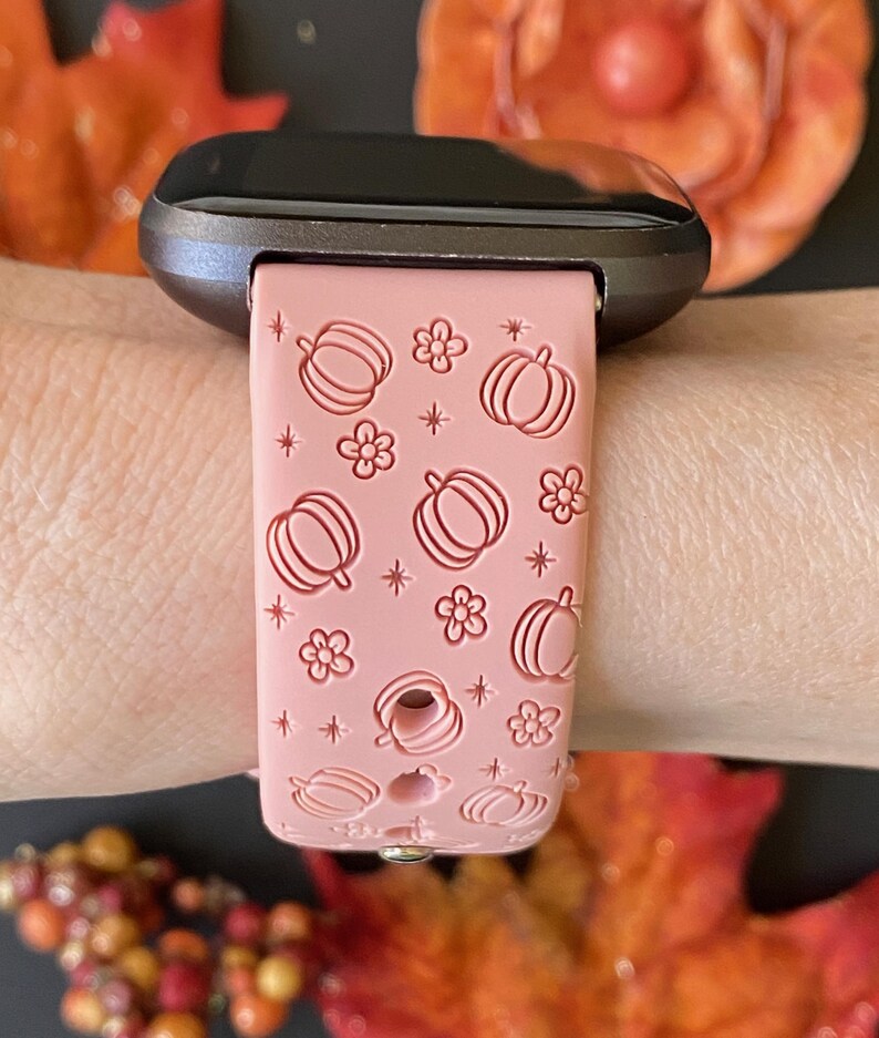 May include: A pink smartwatch band featuring a repeating pattern of pumpkins, flowers, and stars. The band is attached to a dark gray smartwatch face. The design is a seasonal, fall-themed accessory.