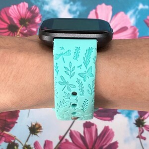 K&ouml;nnte beinhalten: Ein t&uuml;rkisfarbenes Armband f&uuml;r eine Smartwatch mit einem Libellen- und Blattmotiv. Das Armband ist aus Silikon und hat einen schwarzen Metallverschluss.