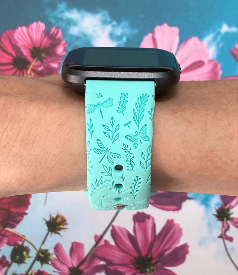 K&ouml;nnte beinhalten: Ein t&uuml;rkisfarbenes Armband f&uuml;r eine Smartwatch mit einem Libellen- und Blattmotiv. Das Armband ist aus Silikon und hat einen schwarzen Metallverschluss.