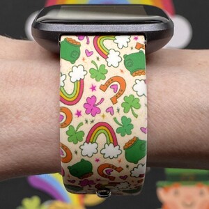 May include: A smartwatch band with a St. Patrick's Day theme. The band features a cream background with colorful rainbows, green pots of gold, horseshoes, and shamrocks. The band is attached to a black smartwatch.