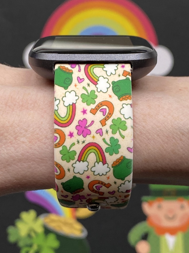 May include: A smartwatch band with a St. Patrick's Day theme. The band features a cream background with colorful rainbows, green pots of gold, horseshoes, and shamrocks. The band is attached to a black smartwatch.