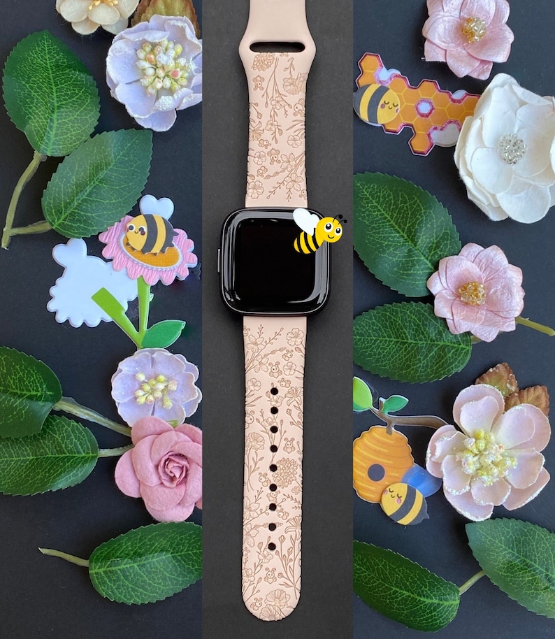 May include: A pink leather watch band with a floral pattern and a black watch face. The band is decorated with small bees and flowers. The watch is surrounded by artificial flowers and leaves.
