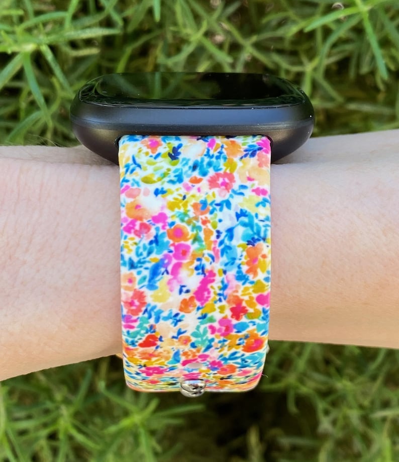 K&ouml;nnte beinhalten: Eine schwarze Smartwatch mit einem bunten, floralen Armband. Das Armband ist aus Silikon und hat einen wei&szlig;en Hintergrund mit kleinen rosa, blauen, gelben und orangefarbenen Blumen.