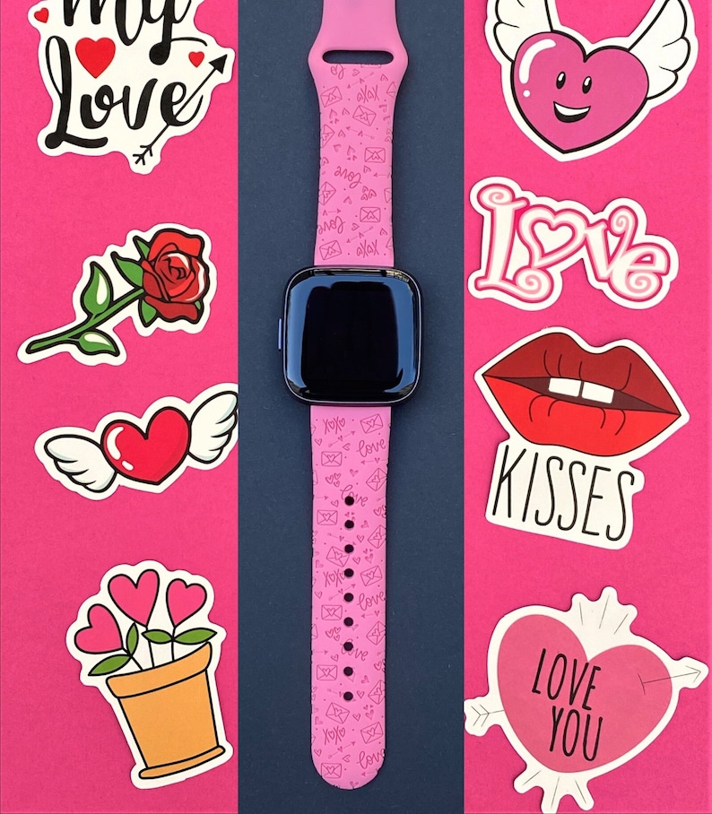 May include: Pink smartwatch with a pink band featuring a repeating pattern of hearts and envelopes. The band has a black buckle. The watch face is black and has a white digital clock.