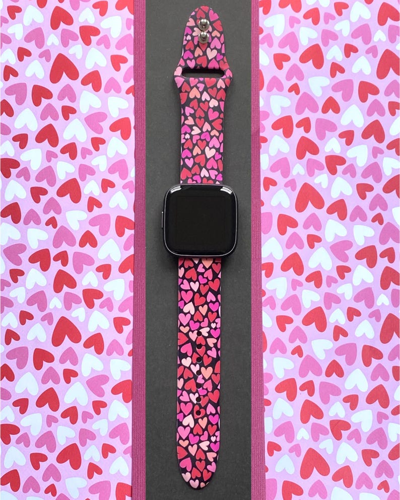 K&ouml;nnte beinhalten: Eine Smartwatch mit schwarzem Bildschirm und einem rosa Armband mit Herzmuster. Das Armband zeigt ein sich wiederholendes Muster aus roten, rosa und wei&szlig;en Herzen auf schwarzem Hintergrund. Die Uhr wird auf einer schwarzen Oberfl&auml;che mit einem rosa Herzmuster-Hintergrund pr&auml;sentiert.