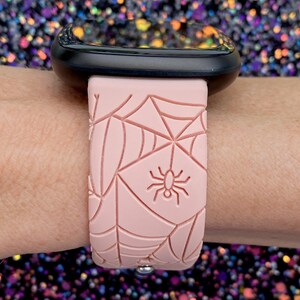 May include: A pink silicone watch band with a spiderweb design and a small spider in the center. The band is designed for a smartwatch.