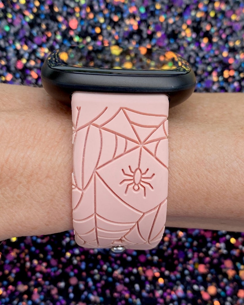 May include: A pink silicone watch band with a spiderweb design and a small spider in the center. The band is designed for a smartwatch.