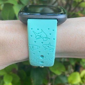 Pode incluir: Uma pulseira de rel&oacute;gio inteligente azul turquesa com um design de tubar&atilde;o e cora&ccedil;&atilde;o. A pulseira &eacute; feita de silicone e tem um mostrador de rel&oacute;gio preto.