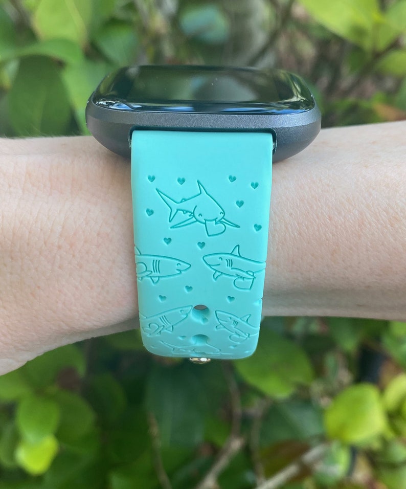 Pode incluir: Uma pulseira de rel&oacute;gio inteligente azul turquesa com um design de tubar&atilde;o e cora&ccedil;&atilde;o. A pulseira &eacute; feita de silicone e tem um mostrador de rel&oacute;gio preto.