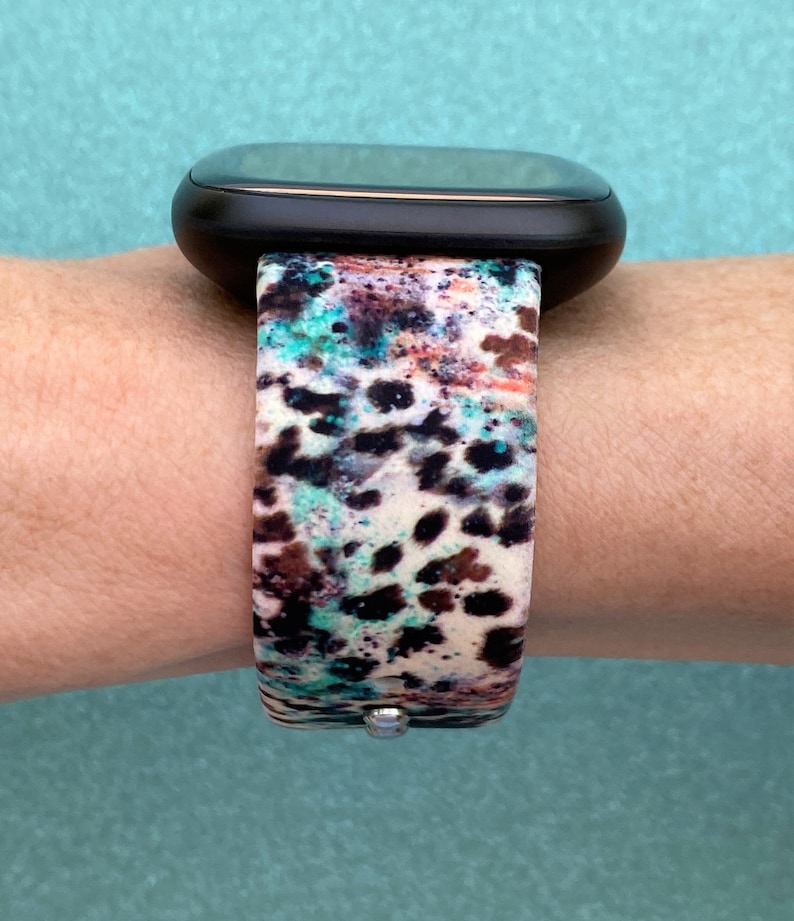 Może przedstawiać: Czarny smartwatch z paskiem w biało-brązowo-niebieskie wzory. Pasek ma wz&oacute;r w panterkę.