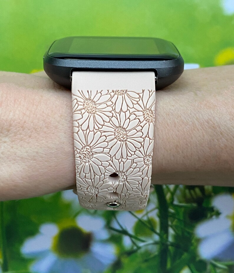 May include: A pink silicone watch band with a daisy pattern. The band is worn on a wrist and has a black watch face.