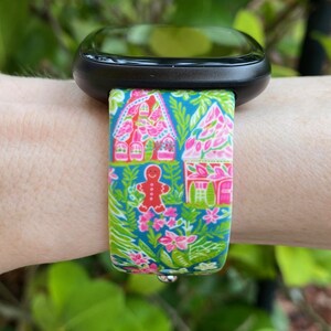 Op de afbeelding: Een zwarte smartwatch met een kleurrijk gingerbreadhuis en een band met een bloemenpatroon. De band is roze, groen en blauw.