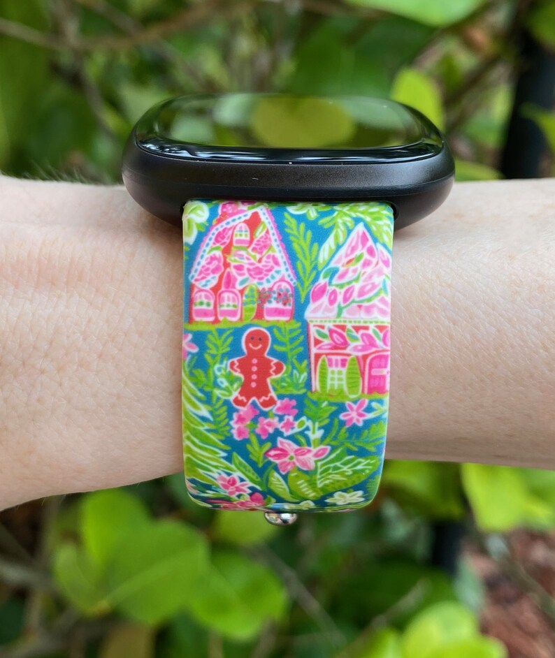 Op de afbeelding: Een zwarte smartwatch met een kleurrijk gingerbreadhuis en een band met een bloemenpatroon. De band is roze, groen en blauw.