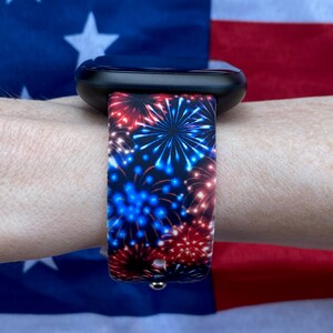 May include: A black smartwatch with a band displaying a firework design in red and blue hues. The watch strap is set against a backdrop of an American flag. The band's pattern is a repeating firework design.