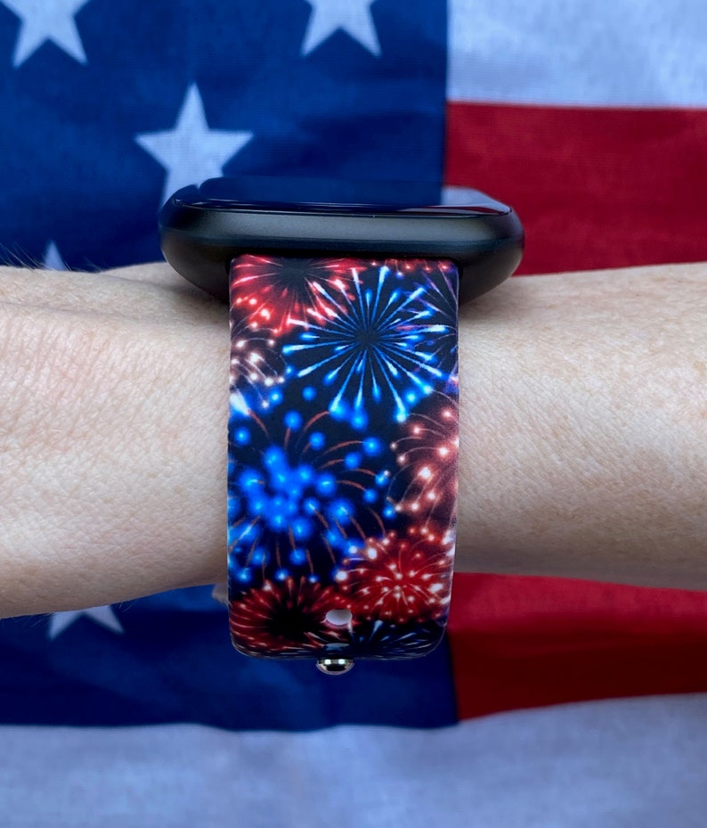 May include: A black smartwatch with a band displaying a firework design in red and blue hues. The watch strap is set against a backdrop of an American flag. The band's pattern is a repeating firework design.