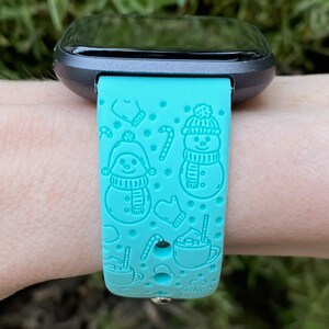 May include: A turquoise blue smartwatch band with a winter holiday theme. The band features a repeating pattern of snowmen, candy canes, mittens, and mugs of hot chocolate.