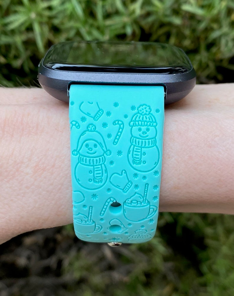 May include: A turquoise blue smartwatch band with a winter holiday theme. The band features a repeating pattern of snowmen, candy canes, mittens, and mugs of hot chocolate.
