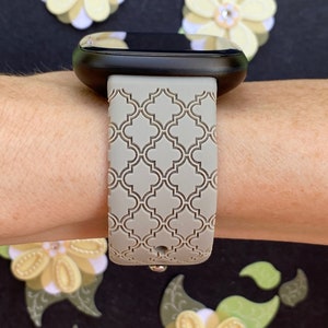 K&ouml;nnte beinhalten: Ein graues, gemustertes Silikon-Armband mit geometrischem Design. Das Armband ist an einer schwarzen Smartwatch befestigt.