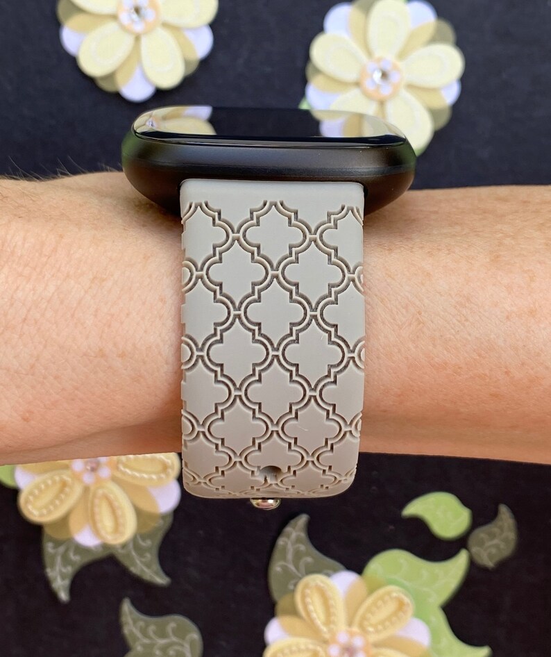K&ouml;nnte beinhalten: Ein graues, gemustertes Silikon-Armband mit geometrischem Design. Das Armband ist an einer schwarzen Smartwatch befestigt.