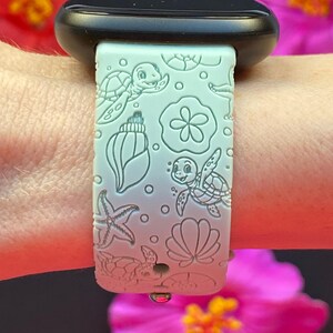 May include: A light green watch band with a sea life design. The band features embossed images of sea turtles, seashells, starfish, and other ocean-themed elements. The watch band is attached to a black watch.