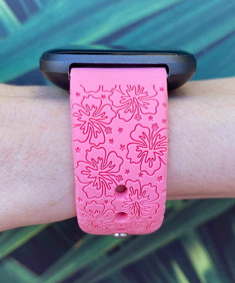 May include: A pink silicone watch band with a hibiscus flower pattern. The band is designed for a smartwatch and has a black watch face.