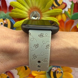 May include: A gray smartwatch with a green bee-themed band. The band has a repeating pattern of bees flying in a circle.