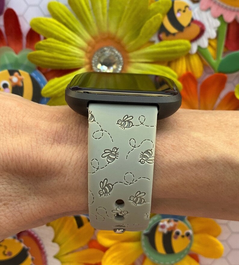 May include: A gray smartwatch with a green bee-themed band. The band has a repeating pattern of bees flying in a circle.