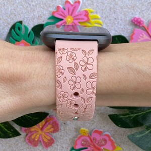 May include: A pink silicone watch band with a floral pattern. The band is designed for a smartwatch and has a black watch face.