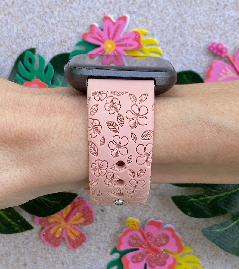 May include: A pink silicone watch band with a floral pattern. The band is designed for a smartwatch and has a black watch face.