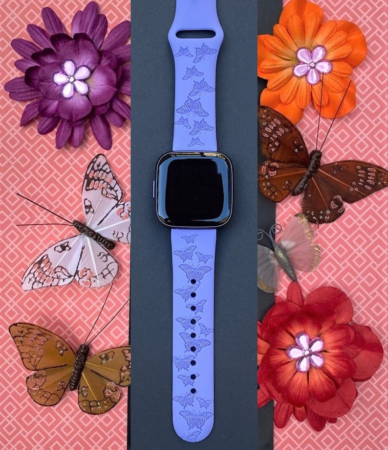 May include: A purple silicone watch band with a butterfly design. The band is on a black background with flowers and butterflies around it.