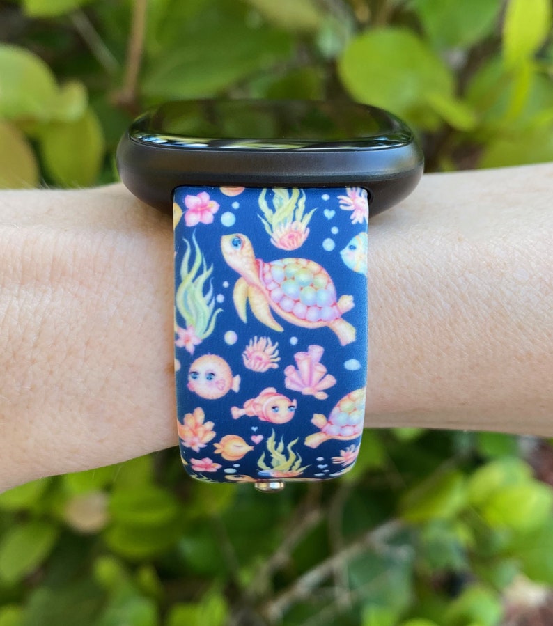 May include: A black smartwatch with a blue and pink patterned band featuring sea turtles, fish, and coral. The band has a colorful, whimsical design.
