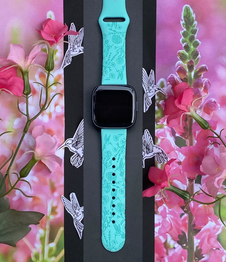 May include: A turquoise smartwatch band with a floral and hummingbird design. The band is made of silicone and has a black watch face.