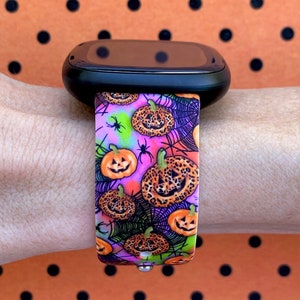 May include: A black smartwatch with a pink and purple patterned band featuring a Halloween theme with pumpkins, spiders, and spiderwebs.