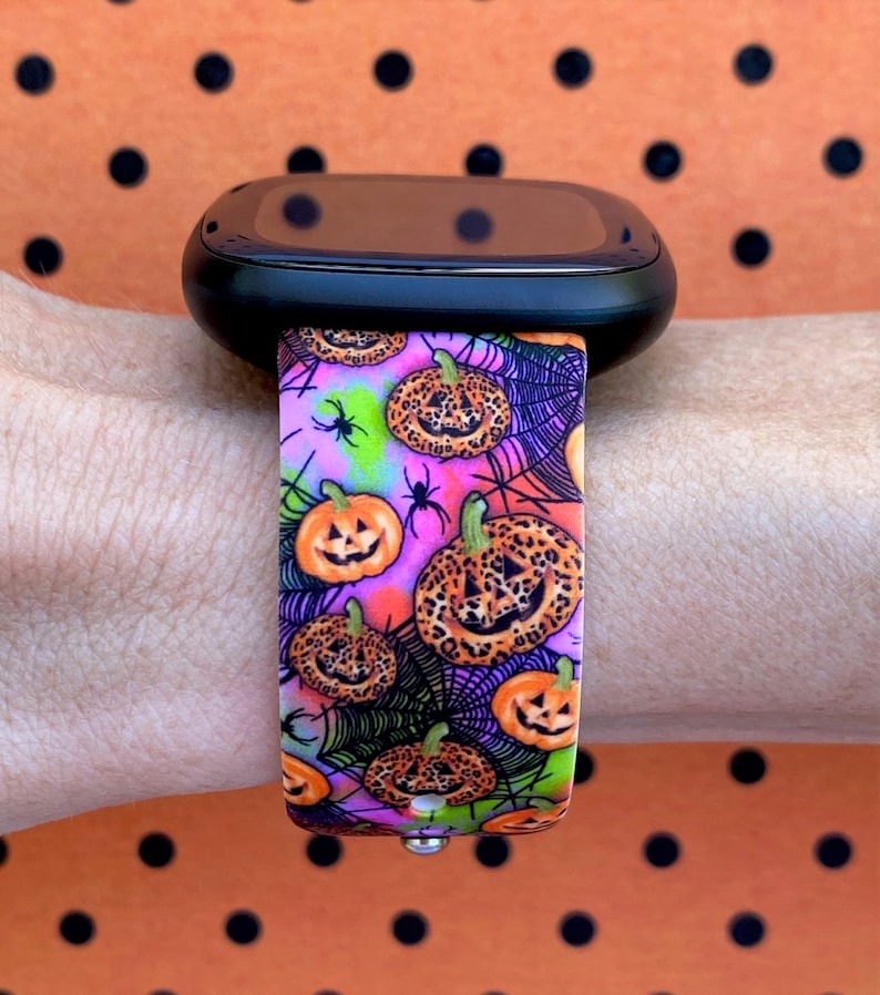May include: A black smartwatch with a pink and purple patterned band featuring a Halloween theme with pumpkins, spiders, and spiderwebs.
