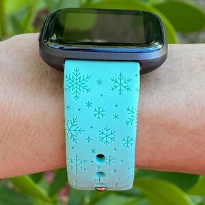 May include: A light blue smartwatch band with a snowflake pattern. The band is made of silicone and has a black metal buckle.