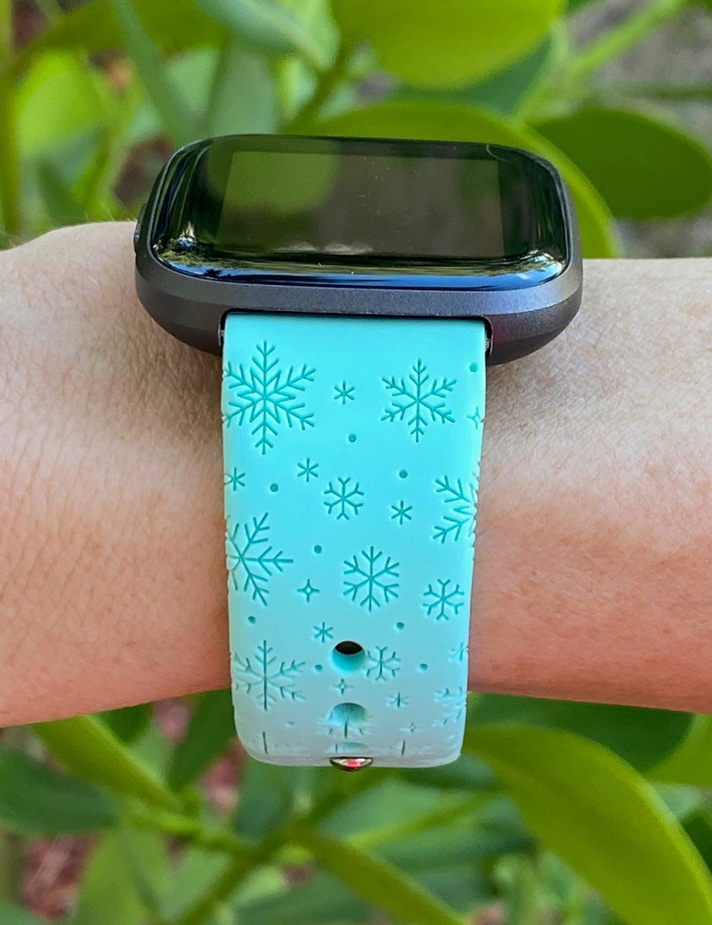 May include: A light blue smartwatch band with a snowflake pattern. The band is made of silicone and has a black metal buckle.