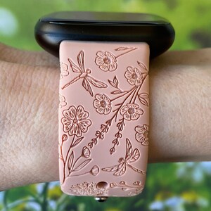 May include: A pink silicone watch band with a floral and dragonfly design. The band is worn on a wrist.