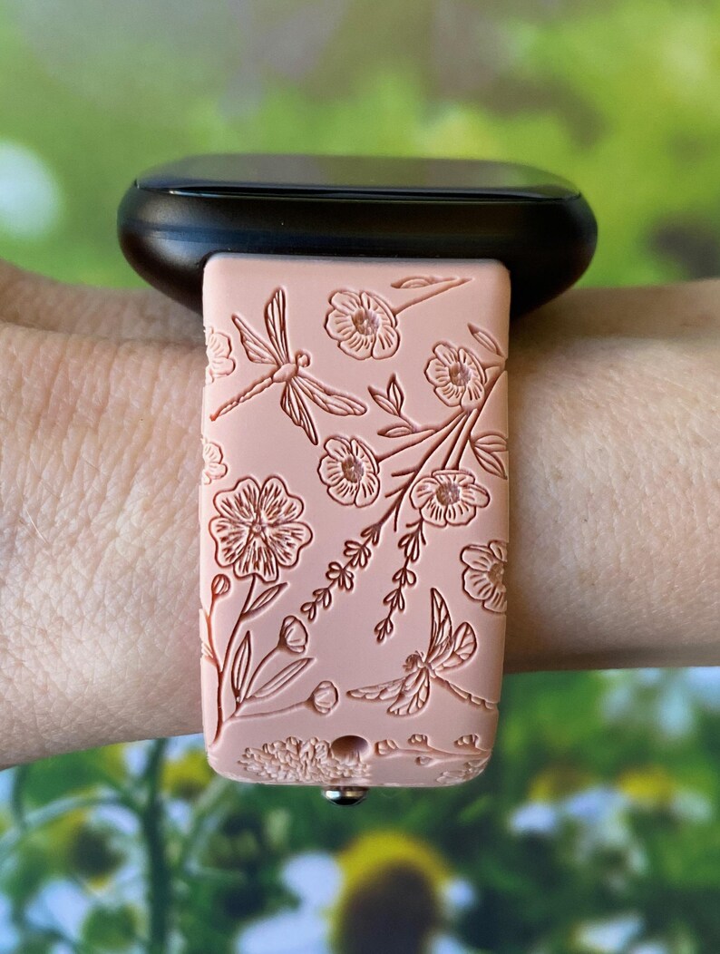 May include: A pink silicone watch band with a floral and dragonfly design. The band is worn on a wrist.