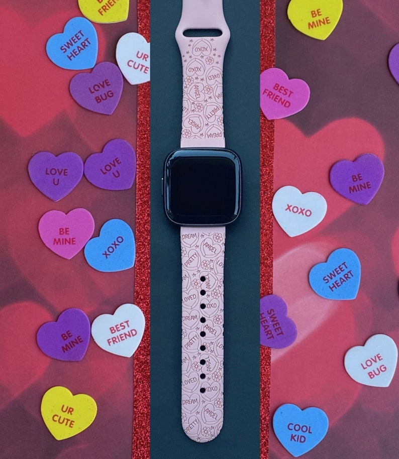 May include: A pink smartwatch strap featuring a pattern of hearts, flowers, and phrases like "Dream" and "Angel." The watch is set amongst colourful conversation heart sweets with messages such as "Sweetheart" and "Love Bug."