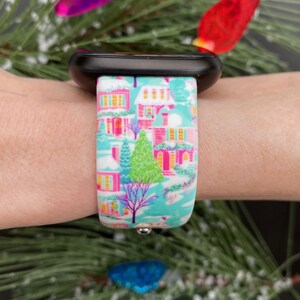 K&ouml;nnte beinhalten: Ein farbenfrohes Uhrenarmband mit einer Winterdorf-Szene mit rosa H&auml;usern, gr&uuml;nen B&auml;umen und einem hellblauen Hintergrund. Das Armband ist an einer schwarzen Smartwatch befestigt. Das Design ist festlich und detailliert, perfekt f&uuml;r die Weihnachtszeit.