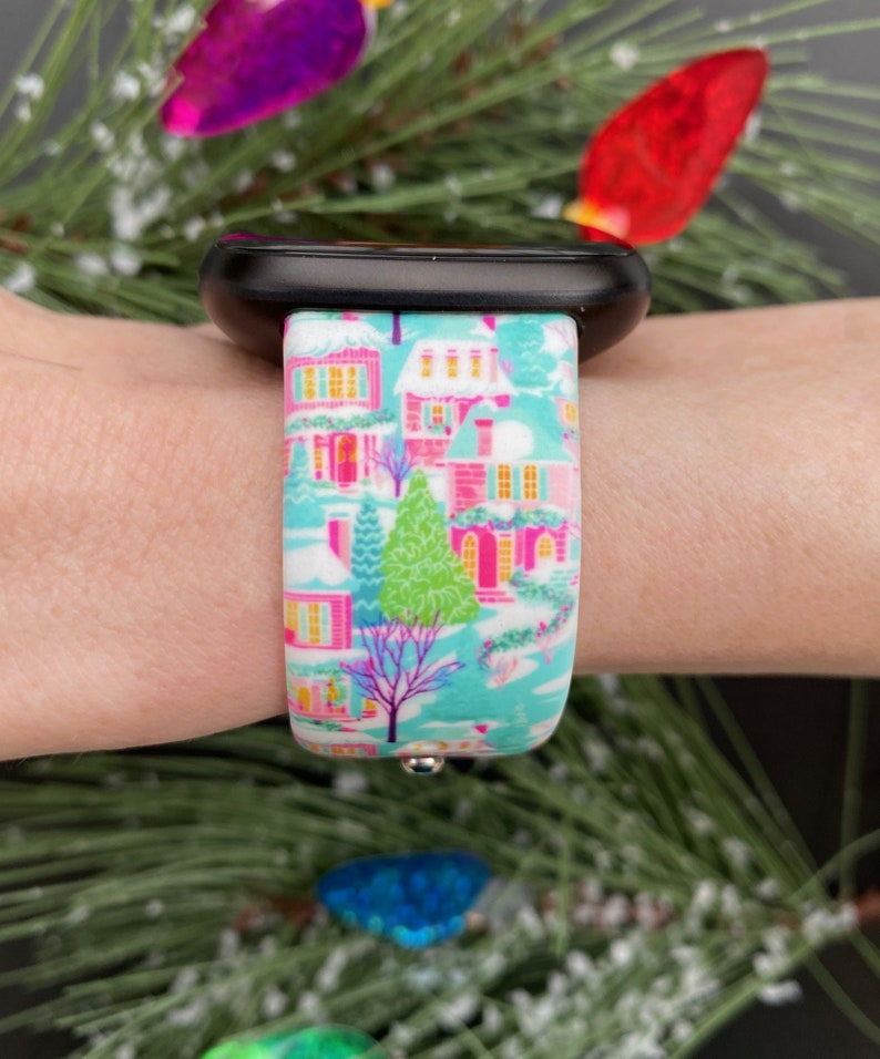 K&ouml;nnte beinhalten: Ein farbenfrohes Uhrenarmband mit einer Winterdorf-Szene mit rosa H&auml;usern, gr&uuml;nen B&auml;umen und einem hellblauen Hintergrund. Das Armband ist an einer schwarzen Smartwatch befestigt. Das Design ist festlich und detailliert, perfekt f&uuml;r die Weihnachtszeit.