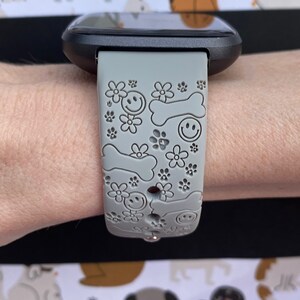 May include: Gray smartwatch band with a pattern of smiling faces, flowers, paw prints, and dog bones. The band is attached to a black smartwatch. The background features a repeating pattern of various dog illustrations.