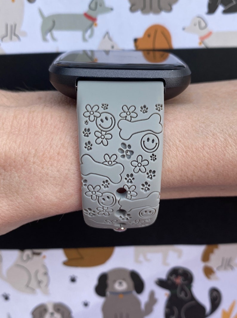 May include: Gray smartwatch band with a pattern of smiling faces, flowers, paw prints, and dog bones. The band is attached to a black smartwatch. The background features a repeating pattern of various dog illustrations.