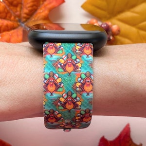 K&ouml;nnte beinhalten: Eine schwarze Smartwatch mit einem bunten Band mit Thanksgiving-Truthahn-Muster. Das Band zeigt ein sich wiederholendes Muster von Truth&auml;hnen in den Farben Rot, Orange, Gelb, Gr&uuml;n und Braun.