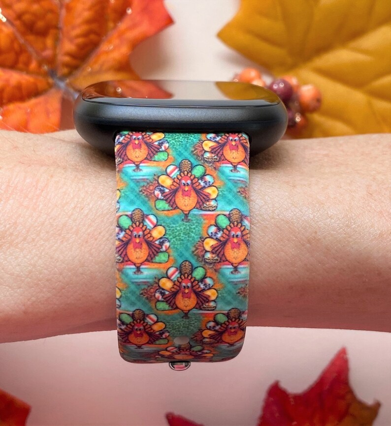 K&ouml;nnte beinhalten: Eine schwarze Smartwatch mit einem bunten Band mit Thanksgiving-Truthahn-Muster. Das Band zeigt ein sich wiederholendes Muster von Truth&auml;hnen in den Farben Rot, Orange, Gelb, Gr&uuml;n und Braun.
