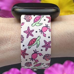 Puede incluir: Correa blanca para smartwatch con un patr&oacute;n repetido de estrellas de mar rosas, cangrejos, tortugas verdes y corales. La correa est&aacute; sujeta a un smartwatch negro. El dise&ntilde;o incluye peque&ntilde;as estrellas negras.