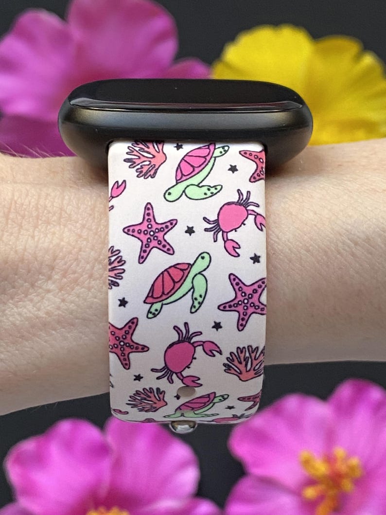Puede incluir: Correa blanca para smartwatch con un patr&oacute;n repetido de estrellas de mar rosas, cangrejos, tortugas verdes y corales. La correa est&aacute; sujeta a un smartwatch negro. El dise&ntilde;o incluye peque&ntilde;as estrellas negras.
