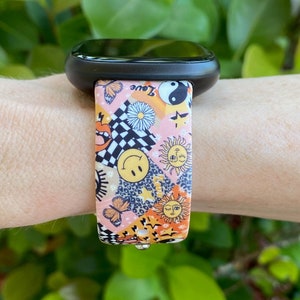 Op de afbeelding: Een roze en oranje gestreept horlogebandje met verschillende ontwerpen, waaronder een smiley, een vlinder, een zon en een yin yang symbool. Het bandje heeft de tekst "Love" erop.